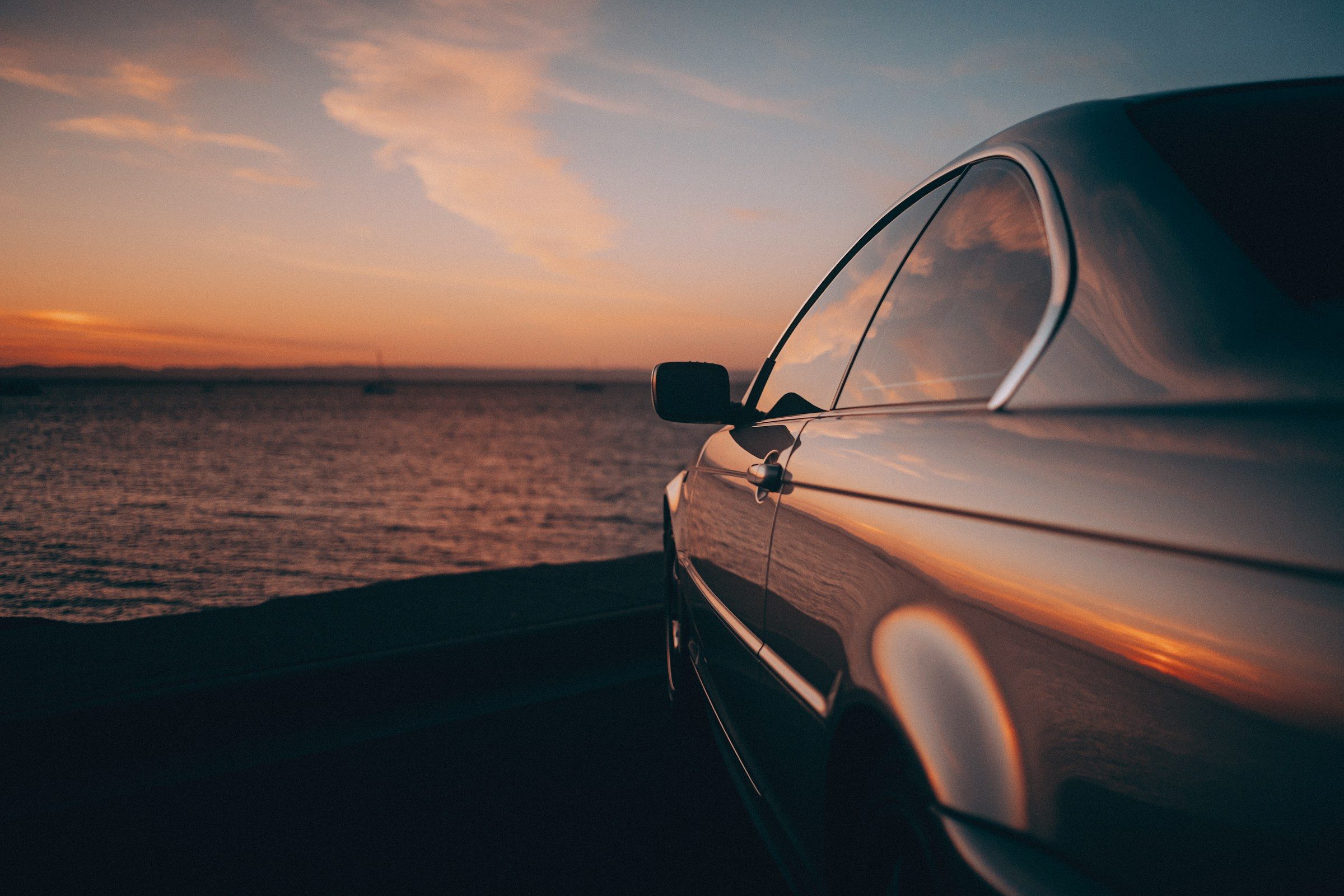 Lateral de um carro escuro estacionado diante do mar ao pôr do sol
